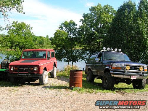 Two Ford Broncos parked outdoors, one red and one blue, with a body of water and trees in the background