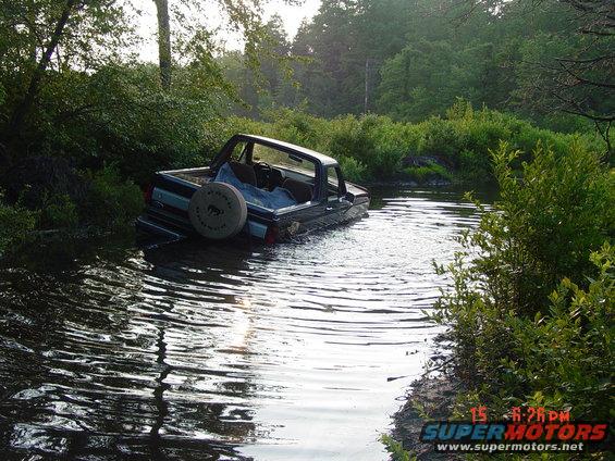 Dark blue pickup truck partially submerged in a stream, spare tire visible