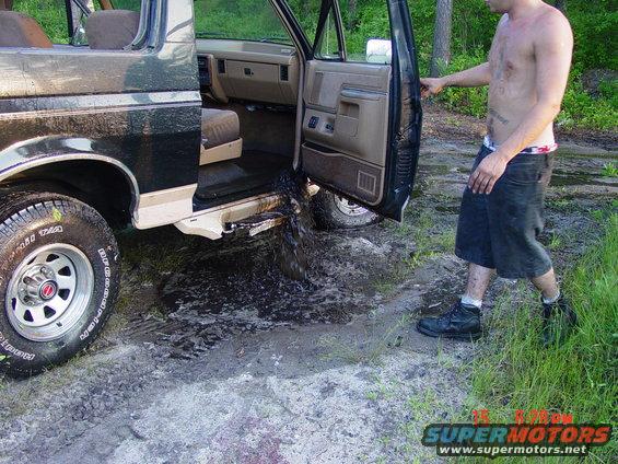 Man standing next to a dark green and tan Ford Bronco with the driver's door open, mud and water pouring out of the interior