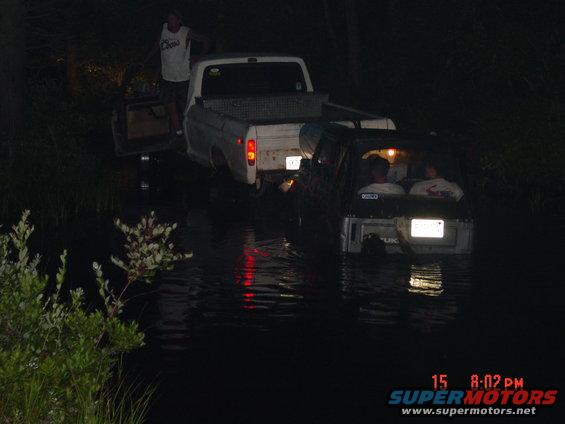 Two trucks are partially submerged in water at night. One truck is white, the other is dark. People are visible inside the dark truck