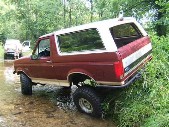 Maroon and white Ford Bronco driving through a shallow stream with tall grass on the bank. Another white SUV is parked nearby