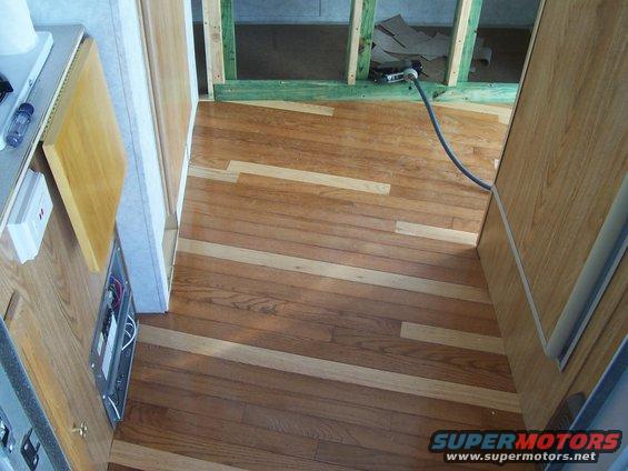 Interior view of a vehicle with wood-paneled walls and unfinished construction, showing hardwood flooring and exposed framing