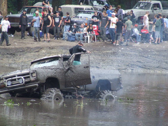 Mud-covered pickup truck stuck in water with a person in the driver's seat. Spectators watch from the bank