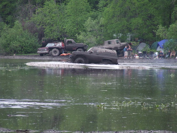 Muddy SUV stuck in water, 3/4 front view. Other trucks and tents visible on muddy bank
