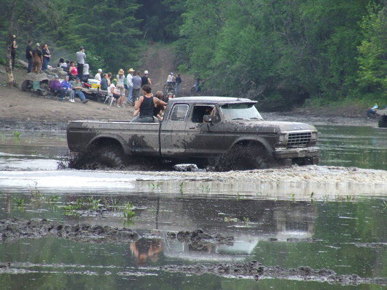 Mud-covered brown Ford pickup truck driving through water, with people watching on the bank
