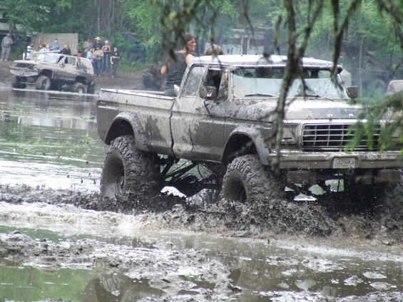 Muddy 1979 Ford F-150 pickup truck with large tires driving through water, another SUV in background