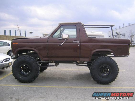 Brown lifted Ford Bronco with large tires, side view. Another car is parked to the left