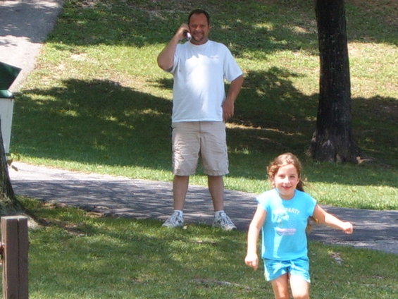 Man talking on cell phone in background, young girl running in foreground on grassy area with trees