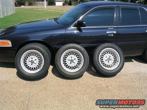 Dark blue sedan, likely a 1998 Ford Crown Victoria, with three aftermarket mesh wheels on the ground in front