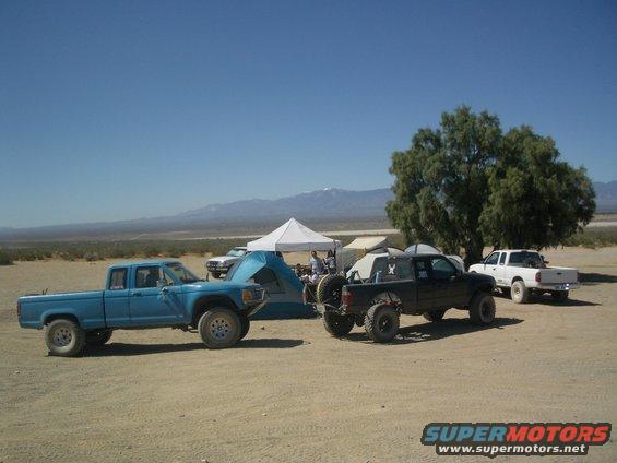 Blue and black off-road trucks parked near tents in a desert landscape with mountains in the background