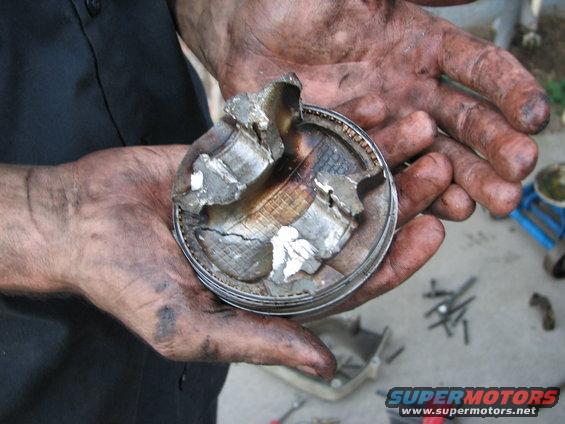 Hands holding a severely damaged engine piston with pieces broken off