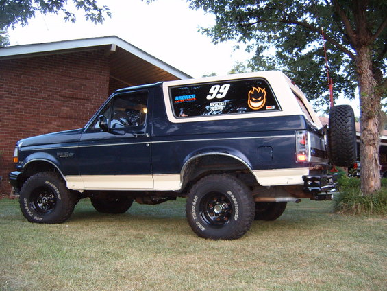 1993 Ford Bronco parked on grass, rear quarter view. Has a spare tire mounted on the back and stickers on the rear window
