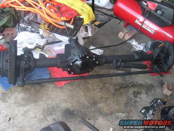 Front axle assembly with differential, brake rotors, and tie rods. Tools and an air compressor are in the background
