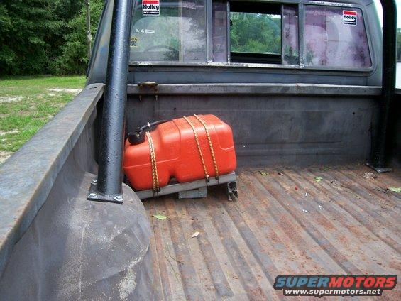 Truck bed interior with orange auxiliary fuel tank secured by ropes. Holley stickers visible on rear window