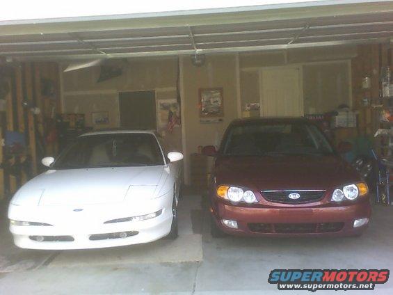 White car and red car parked in a garage. The red car has a Kia logo