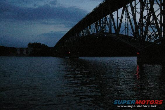 Large steel bridge over dark water at dusk. Buildings visible on the far shore