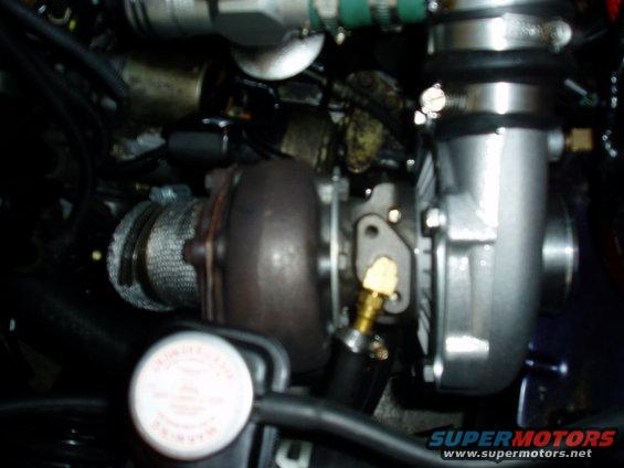 Turbocharger with heat wrap and silver housing, connected to engine components. A white cap with red text is visible