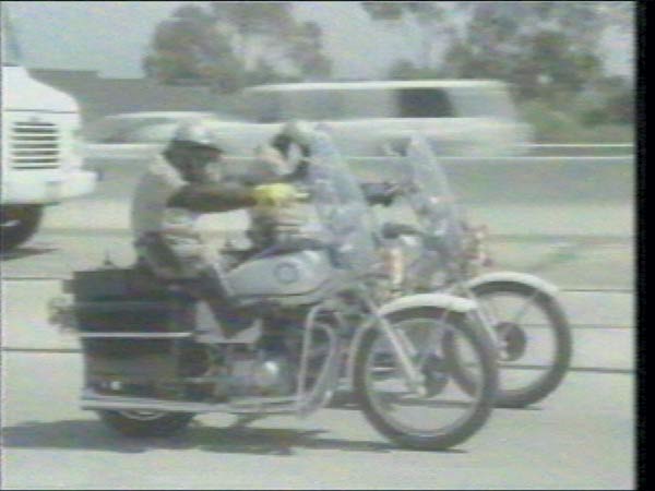 Two motorcycles with riders on a road, one with a large cargo box behind the seat