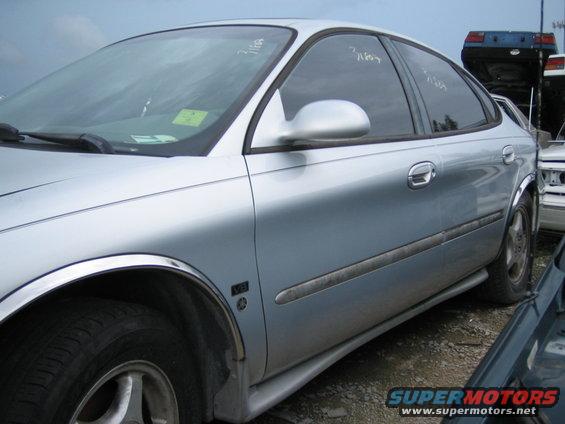 Silver sedan with chrome trim, parked in a junkyard. Another car is visible in the foreground, and a blue vehicle is in the background