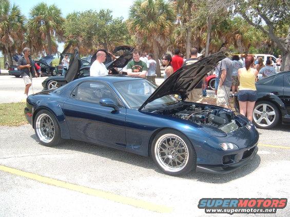 Blue Mazda RX-7 with hood open, showing engine. People gathered around at an outdoor car show