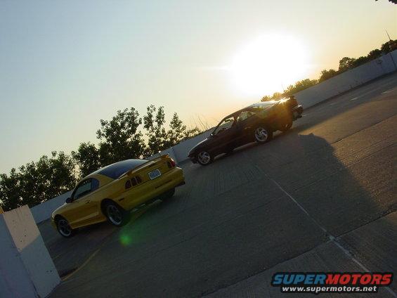 Yellow 1998 Ford Mustang and dark car on highway at sunset