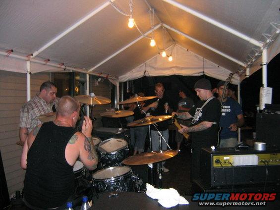 Band performing under a white tent with string lights, drummer in foreground