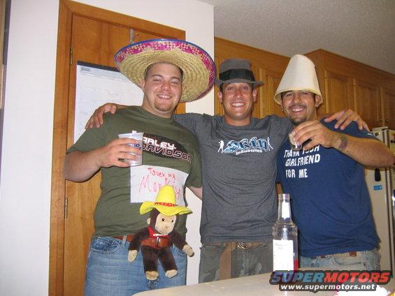 Three men posing for a photo, two wearing novelty hats. One man has a monkey toy attached to his shirt