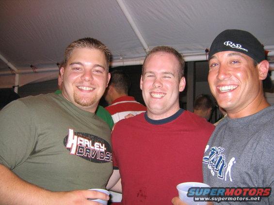 Three smiling men at an outdoor gathering, one wearing a Harley Davidson shirt