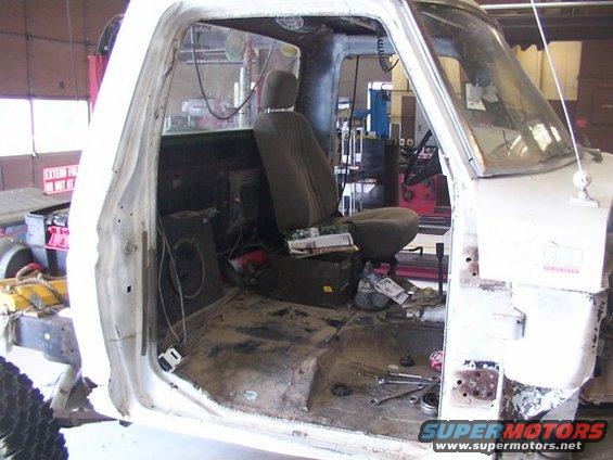 Interior of a white pickup truck, driver's side door removed, showing the seat and floor in disarray. Tools are on the floor