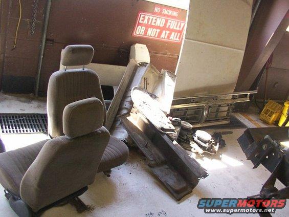 Two car seats and various vehicle parts are piled on a garage floor. A sign reads 'NO SMOKING EXTEND FULLY OR NOT AT ALL'