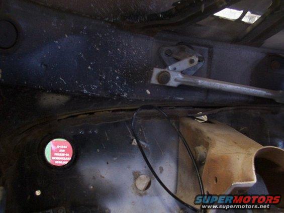 Underside of a vehicle showing the wiper linkage and a red circular sticker with text