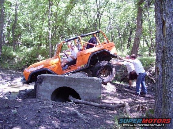 Orange off-road vehicle with roll cage going over concrete obstacle, two people pushing