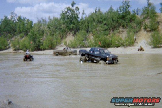 Black pickup truck stuck in mud, with ATVs and another truck in the background