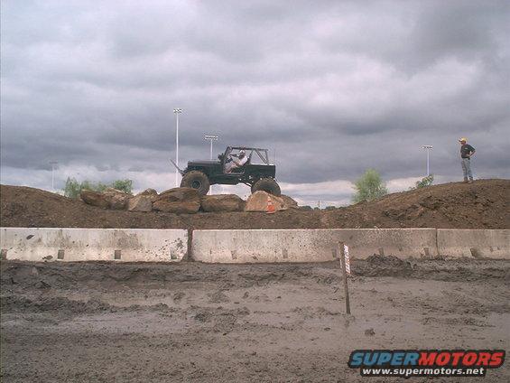 Jeep climbing over rocks on a dirt mound, with a person watching from a higher hill