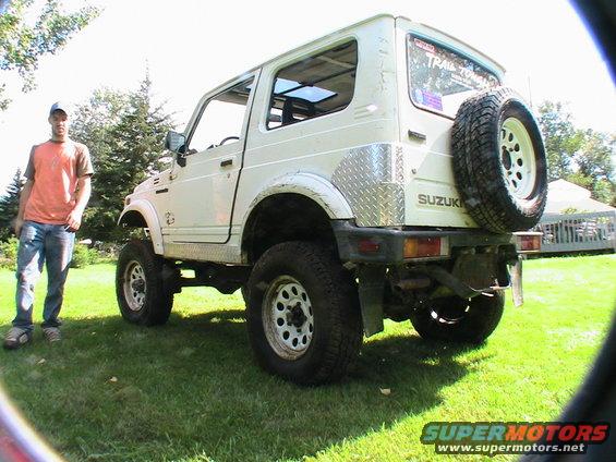 White Suzuki Samurai parked on grass with a person standing to its left. Spare tire mounted on the rear