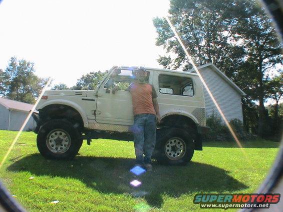 Man leaning on a white Suzuki Samurai with large tires parked on grass in front of a house