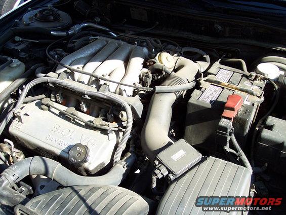 Engine bay with "3.0L V6 24 VALVE" text visible on a silver cover, a black battery, and various hoses and wires