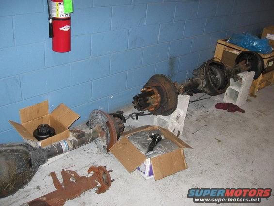 Several rusty rear axles and differential parts are laid out on a concrete floor, with a fire extinguisher mounted on a blue brick wall