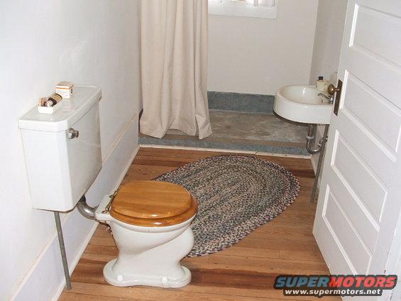 Bathroom with a white toilet, wooden seat, and a woven rug on a wooden floor