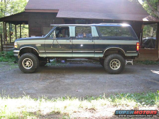 Lifted Ford Bronco SUV, dark blue and light blue two-tone paint, parked in front of a wooden cabin