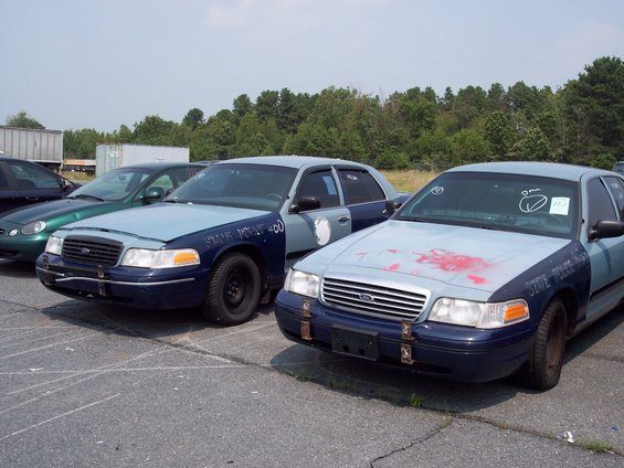Two police cars, light blue and dark blue, parked in a lot. One has red spray paint on the hood