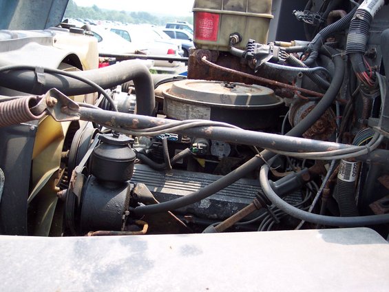Engine compartment with hoses, wires, and engine block visible. Some rust is present