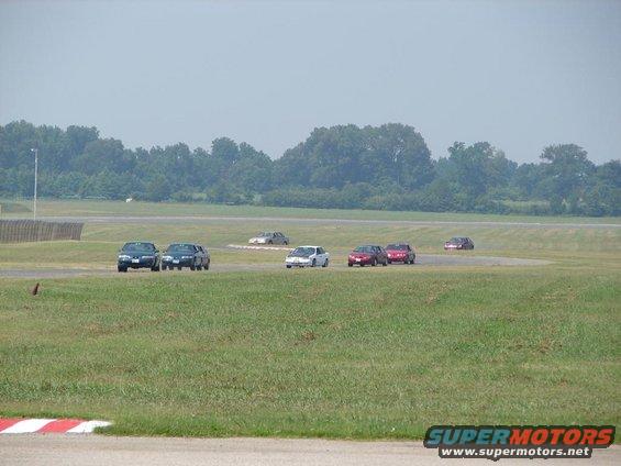 Several sedans race on a track, with trees in the background
