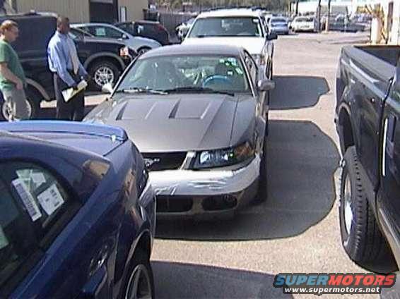 Front view of a silver Ford Mustang with hood scoops and tape on the bumper. A blue car is in the foreground