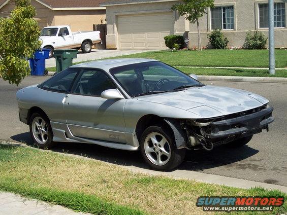 Silver coupe parked on street, front bumper missing, showing exposed engine components. White pickup truck in driveway behind it