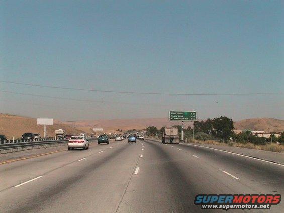 Highway traffic under a clear blue sky with distant hills and a green road sign
