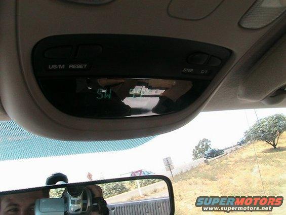 Overhead console with digital display showing '5W' and buttons labeled 'US/M RESET' and 'STEP C/T'. Reflection shows a person holding a video camera