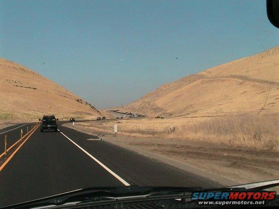 Black SUV driving on a highway through a valley with dry grass and hills