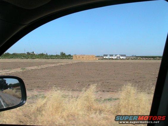 View from car window of a rural field with hay bales and a white house under a clear blue sky