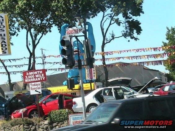 Car dealership lot with multiple cars, a large blue monster figure, and various signs